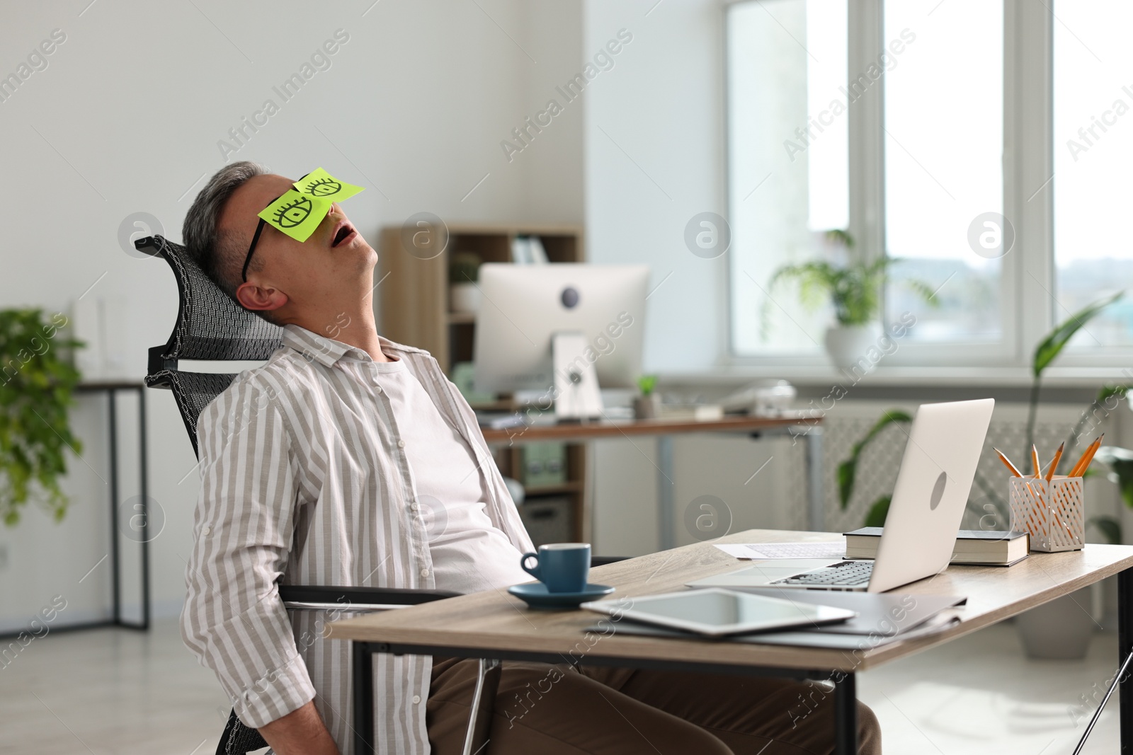 Photo of Man with fake eyes painted on sticky notes snoozing at workplace in office