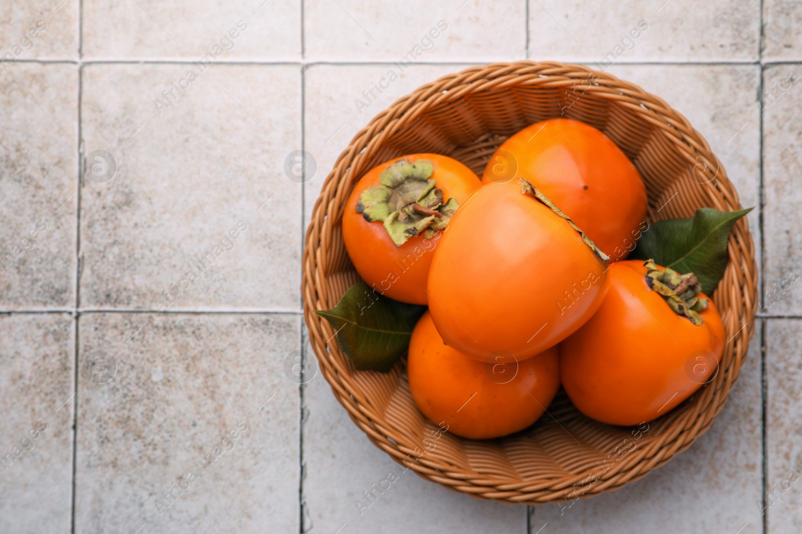 Photo of Delicious ripe juicy persimmons in wicker basket on tiled surface, top view. Space for text