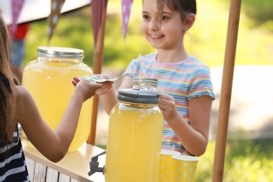 Photo of Little girl selling natural lemonade to kid in park. Summer refreshing drink