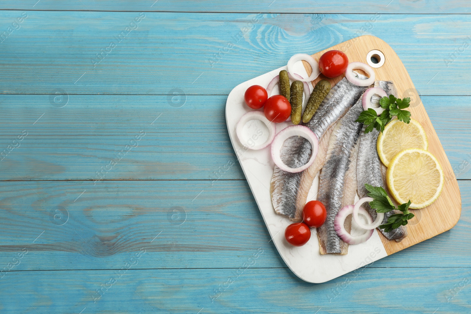 Photo of Serving board with salted herring fillets, parsley, onion rings, pickles, cherry tomatoes and lemon on light blue wooden table, top view. Space for text