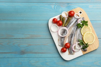 Serving board with salted herring fillets, parsley, onion rings, pickles, cherry tomatoes and lemon on light blue wooden table, top view. Space for text
