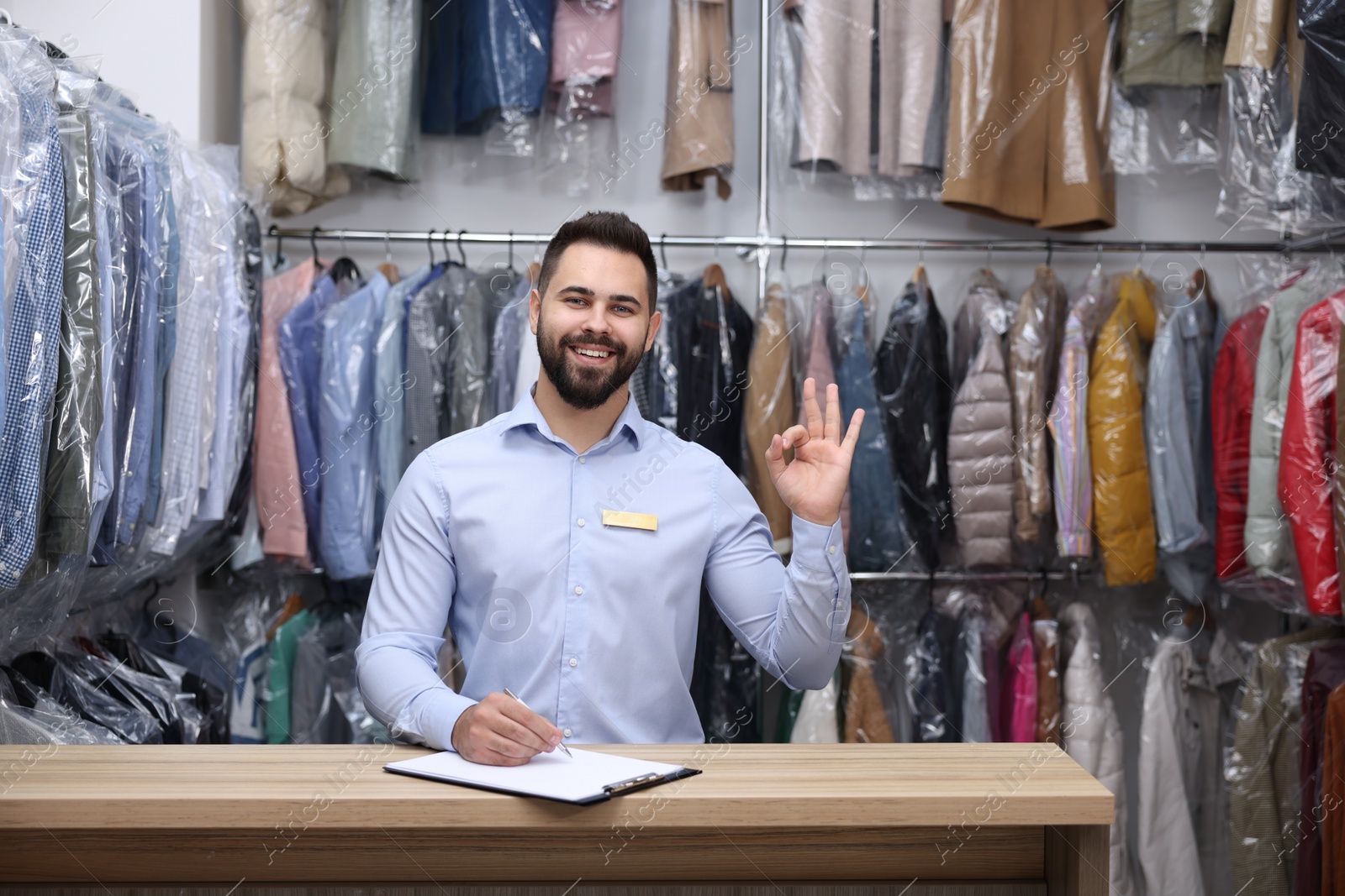 Photo of Dry-cleaning service. Happy worker showing ok gesture at counter indoors