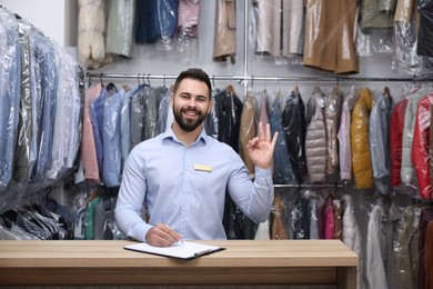 Dry-cleaning service. Happy worker showing ok gesture at counter indoors