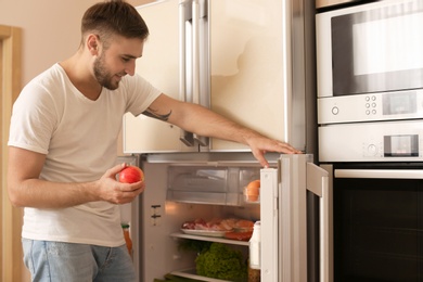 Photo of Young man taking apple from refrigerator in kitchen