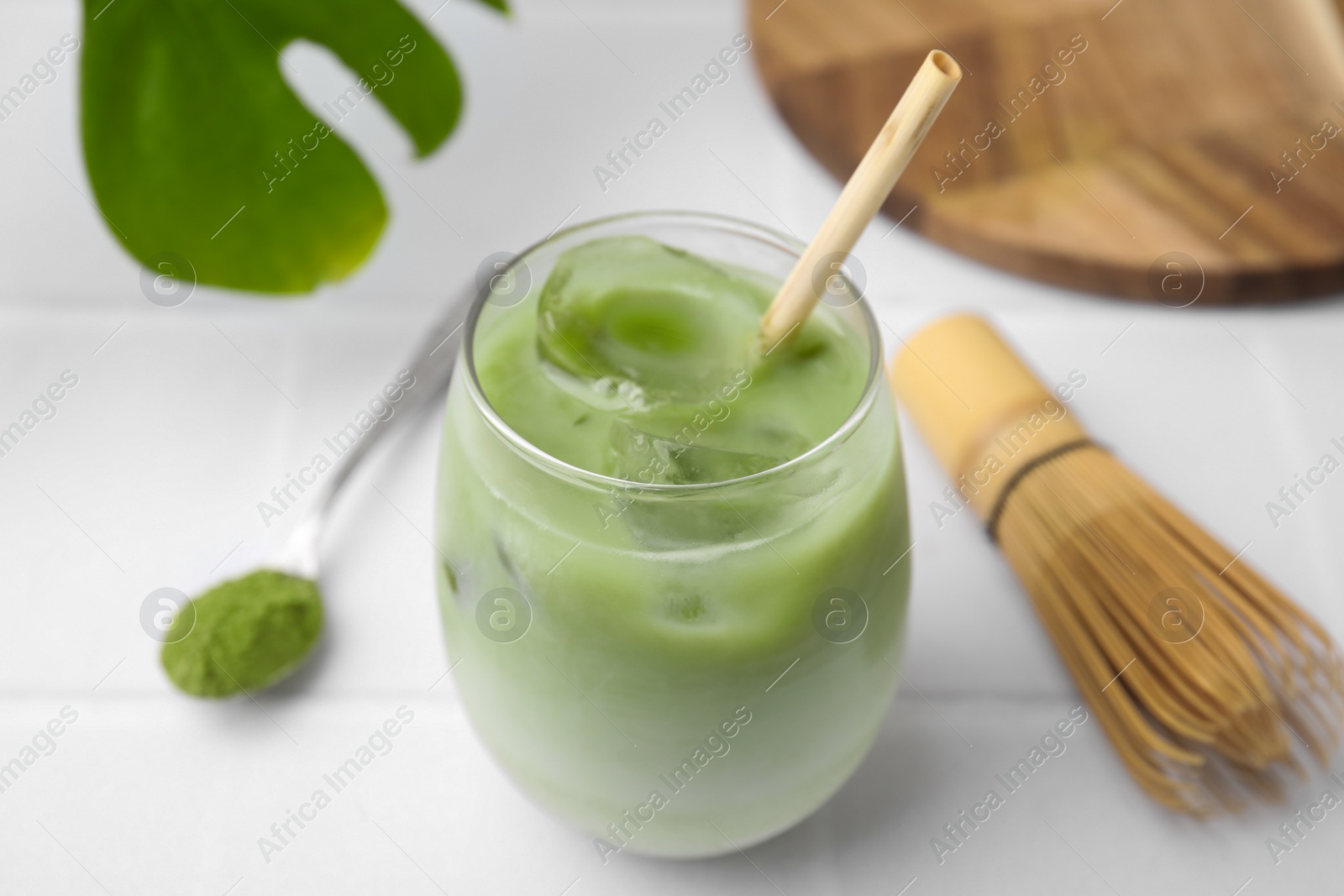Photo of Glass of tasty iced matcha latte, leaf, bamboo whisk and powder on white tiled table, closeup