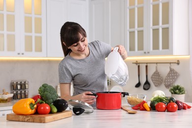 Happy young housewife pouring water in pot at white marble table in kitchen. Cooking process