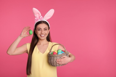 Photo of Beautiful woman in bunny ears headband holding basket with Easter eggs on color background, space for text