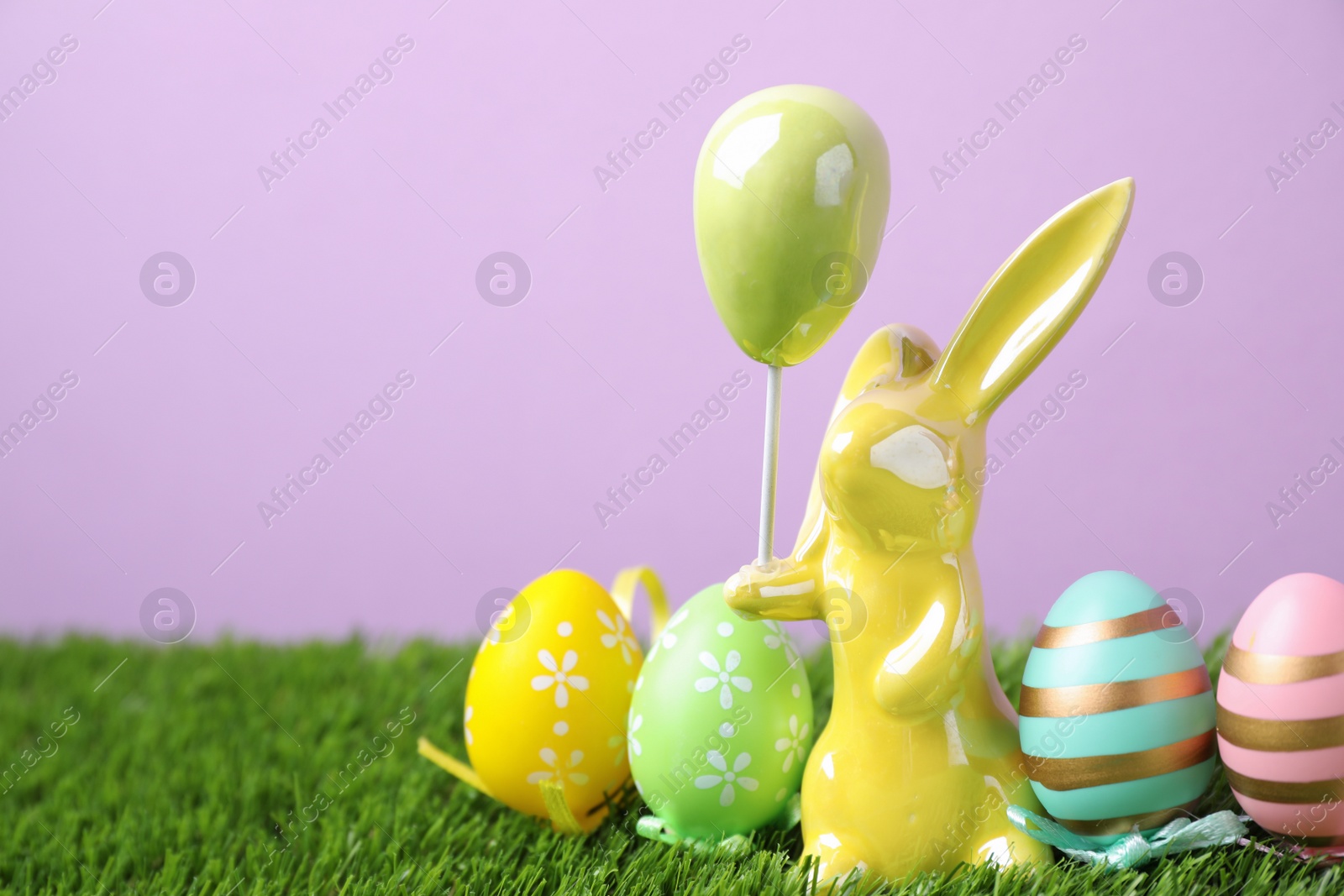 Photo of Easter bunny figure and dyed eggs on green grass against violet background. Space for text