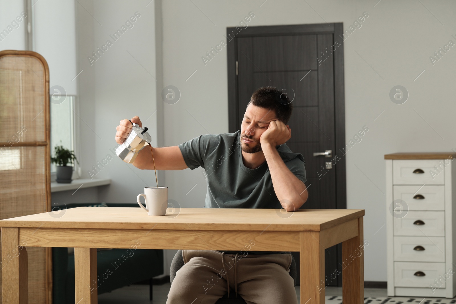 Photo of Sleepy man pouring coffee in cup at wooden table indoors