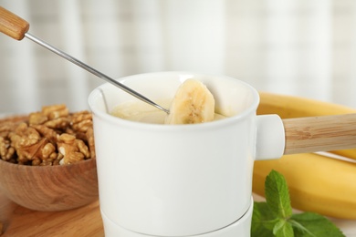 Dipping slice of banana into fondue pot with white chocolate indoors, closeup