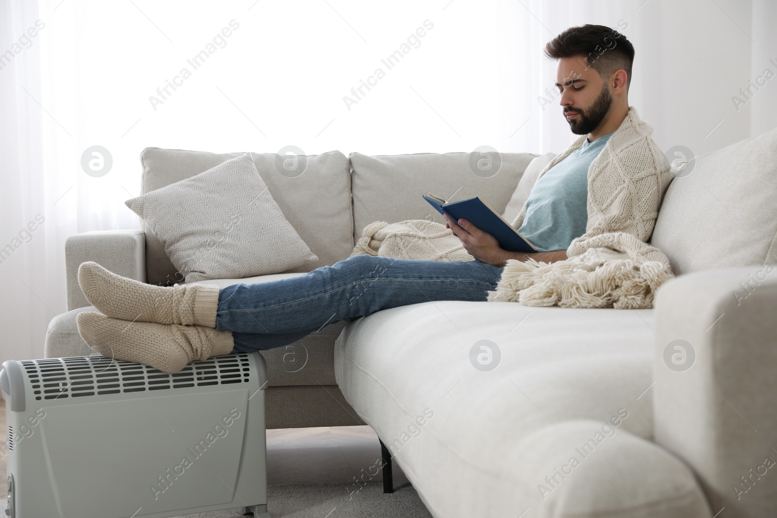 Photo of Young man with book warming feet on electric heater at home