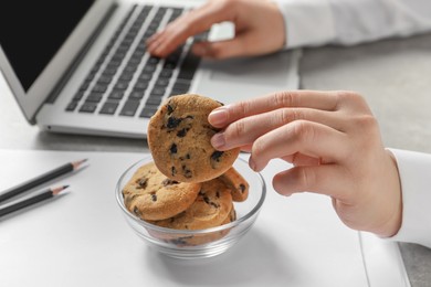 Woman taking chocolate chip cookie from bowl while working with laptop in office, closeup