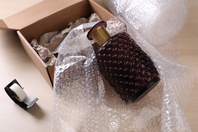 Beautiful glass vase in bubble wrap near cardboard box with paper and adhesive tape on wooden table