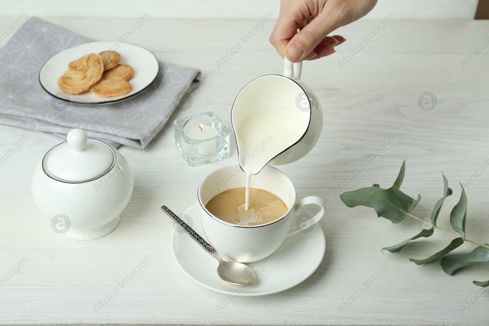 Photo of Woman pouring milk into cup with coffee at white wooden table, closeup