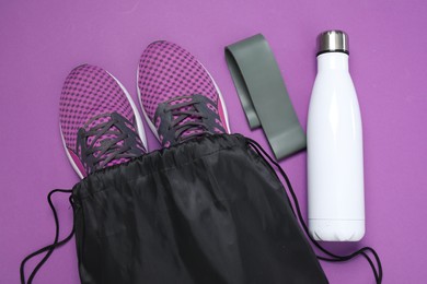 Photo of Black drawstring bag, sneakers, thermo bottle and fitness elastic band on purple background, flat lay