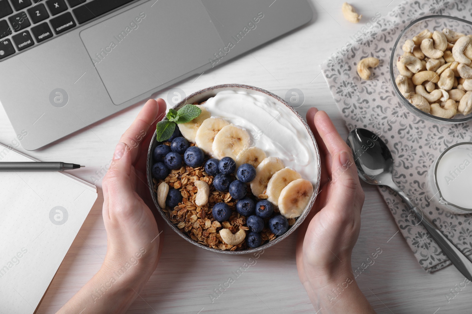 Photo of Woman holding bowl of tasty granola at white wooden table with laptop, top view