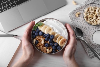 Photo of Woman holding bowl of tasty granola at white wooden table with laptop, top view