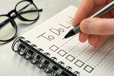 Photo of Woman filling To Do list in notepad at light grey table, closeup