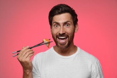 Emotional man holding tasty sushi with chopsticks on pink background