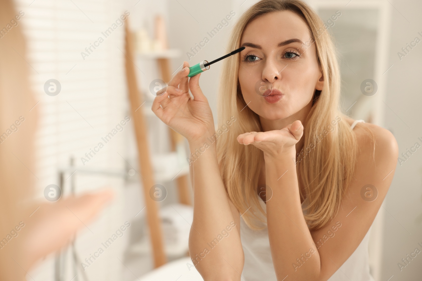 Photo of Beautiful woman blowing kiss while applying mascara near mirror in bathroom