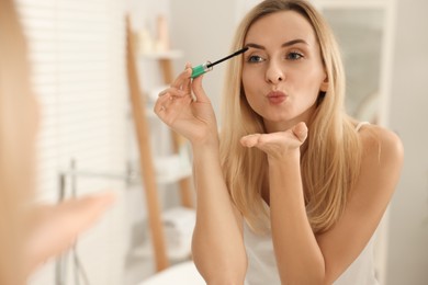 Photo of Beautiful woman blowing kiss while applying mascara near mirror in bathroom