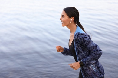 Photo of Young sportswoman with wireless earphones running near river