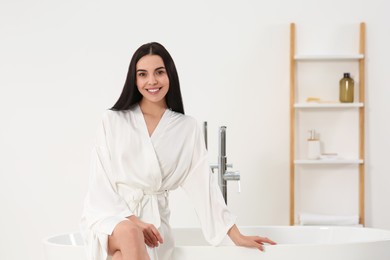 Beautiful happy woman in stylish bathrobe sitting on tub in bathroom