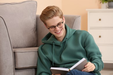 Photo of Teenage boy reading book near armchair in room