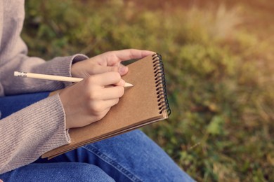 Photo of Young woman drawing with pencil in sketchbook outdoors, closeup