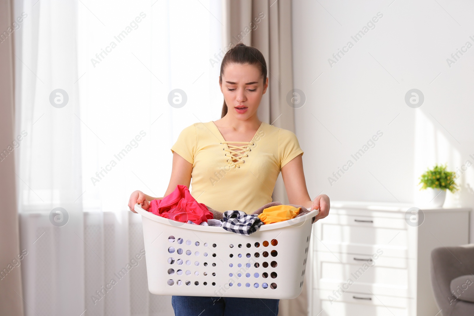 Photo of Displeased young woman holding basket with laundry at home
