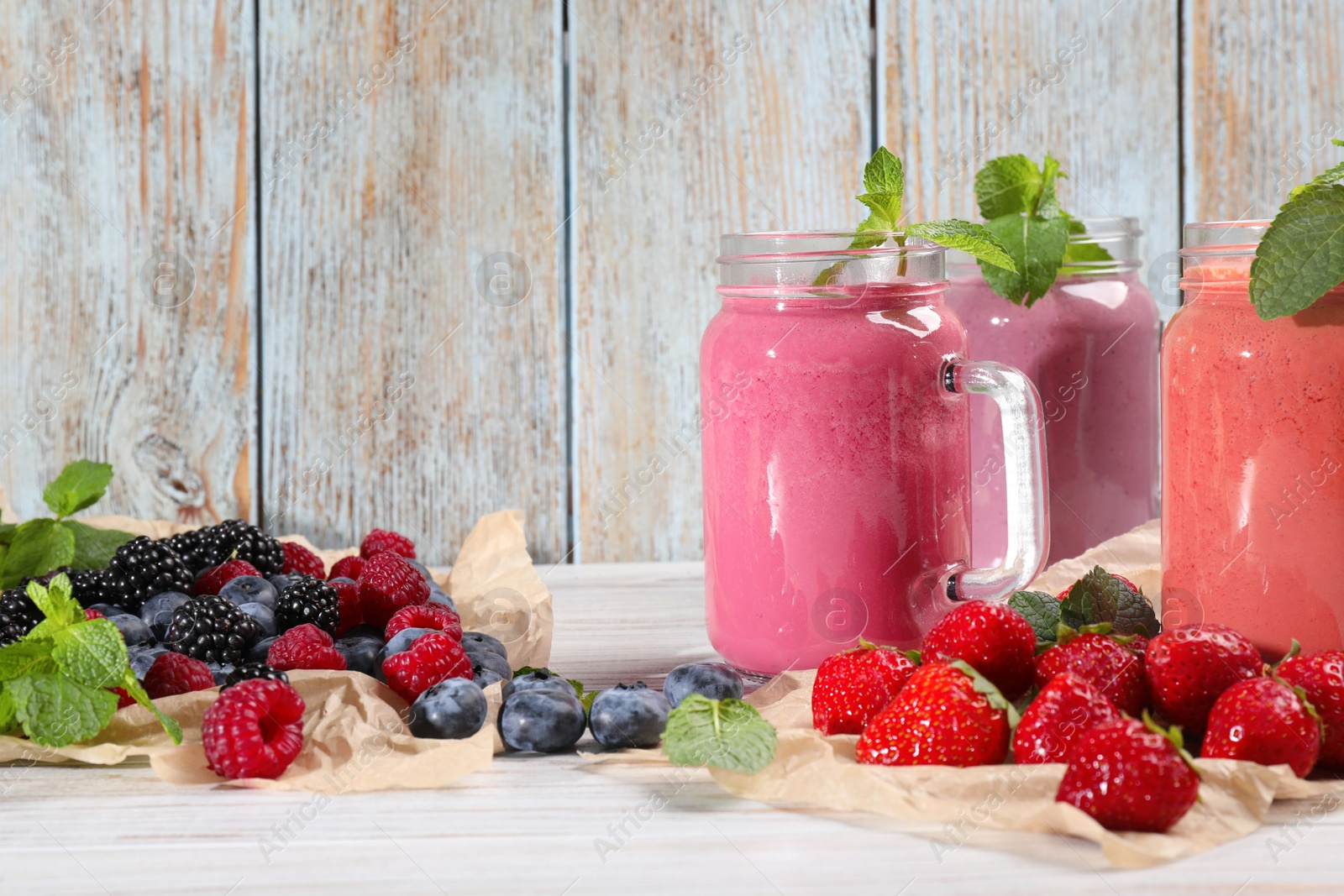 Photo of Mason jars of different berry smoothies and fresh ingredients on white wooden table