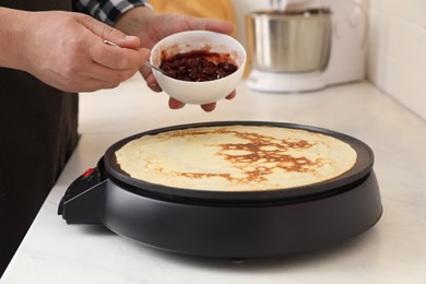 Photo of Man cooking delicious crepe with jam on electric pancake maker in kitchen, closeup