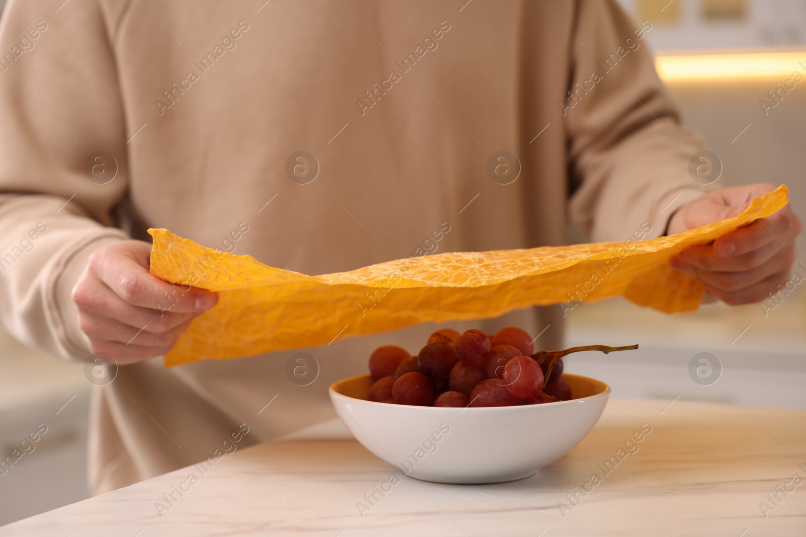 Photo of Man packing bowl of fresh grapes into beeswax food wrap at light marble table in kitchen, closeup