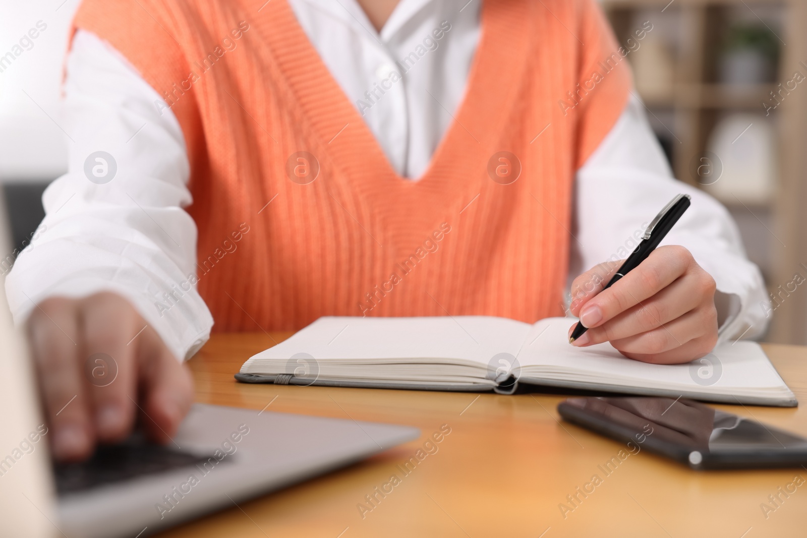 Photo of Young woman writing in notebook while working on laptop at wooden table, closeup