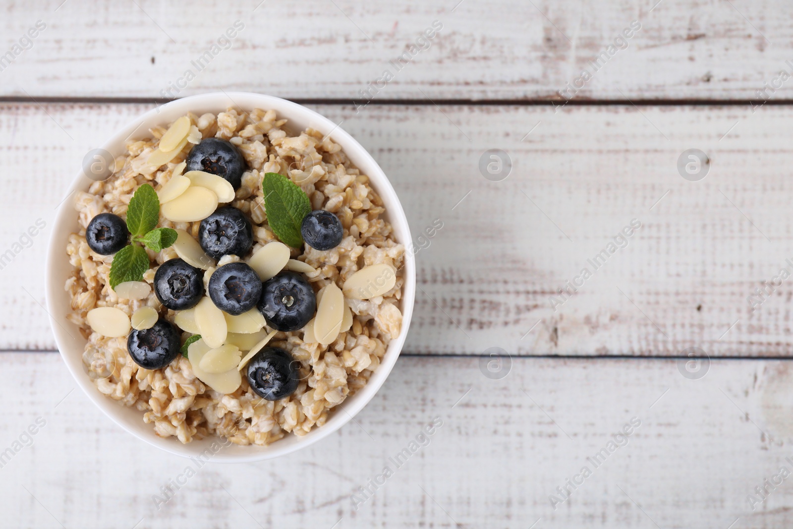 Photo of Tasty oatmeal with blueberries, mint and almond petals in bowl on white wooden table, top view. Space for text