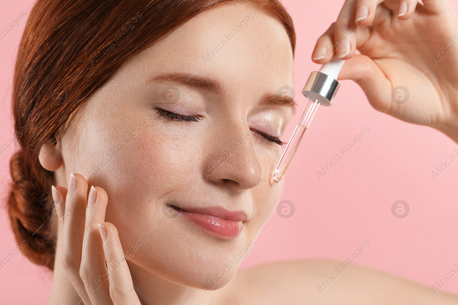 Photo of Beautiful woman with freckles applying cosmetic serum onto her face on pink background, closeup