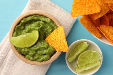 Photo of Bowl of delicious guacamole, lime and nachos chips on light blue wooden table, flat lay
