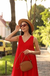Photo of Beautiful young woman with stylish bag in red dress and sunglasses outdoors