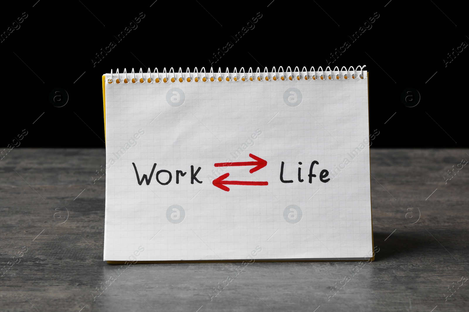 Photo of Notebook with words Work, Life and arrows on grey table against black background. Balance concept