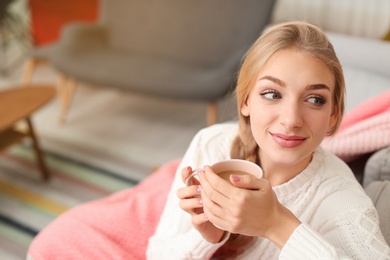 Photo of Beautiful young woman wrapped in plaid sitting on sofa with cup at home. Winter atmosphere