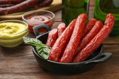Photo of Thin dry smoked sausages, basil and sauces on wooden table, closeup