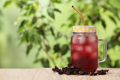 Refreshing hibiscus tea with ice cubes in mason jar and dry roselle flowers on wooden table against blurred green background. Space for text