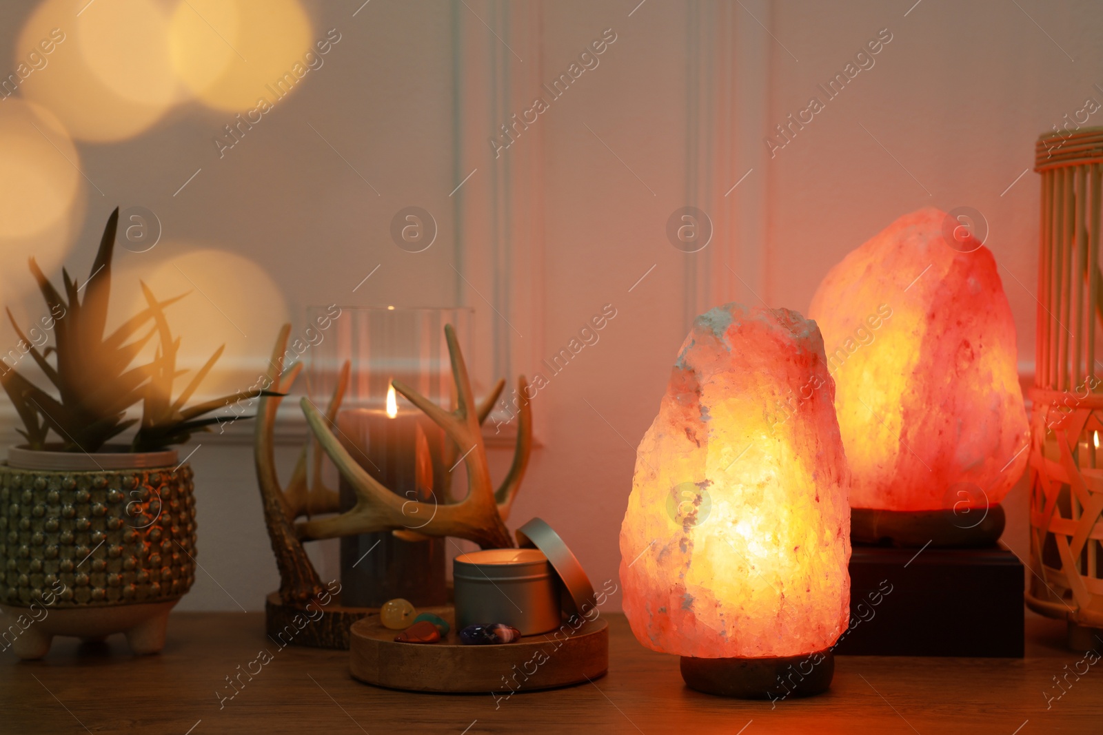 Photo of Himalayan salt lamps, candles, houseplant and gemstones on wooden table near white wall indoors. Bokeh effect