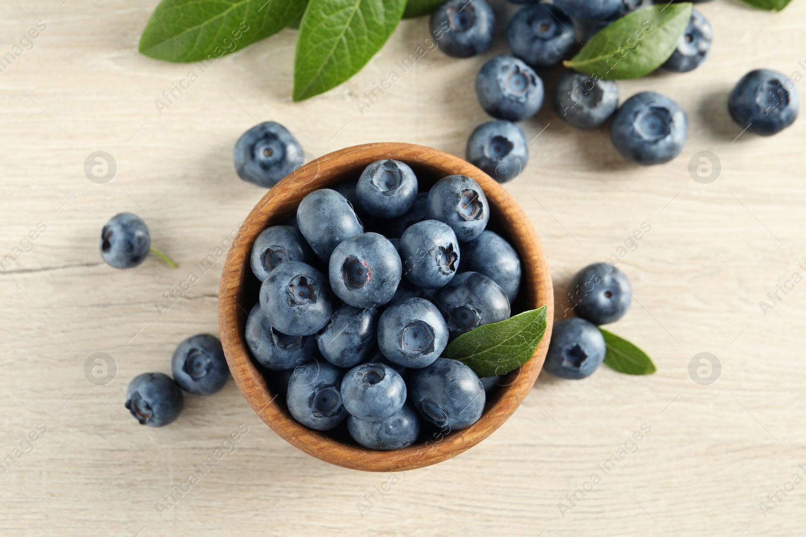 Photo of Bowl of fresh tasty blueberries with leaves on white wooden table, flat lay