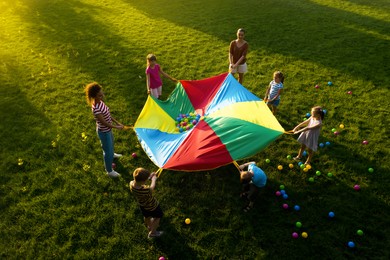 Group of children and teachers playing with rainbow playground parachute on green grass, above view. Summer camp activity
