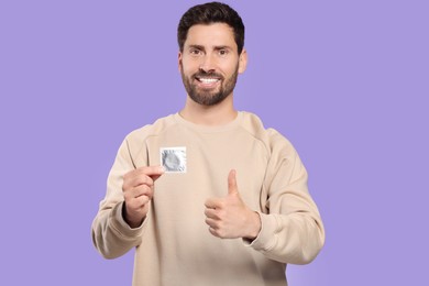 Happy man with condom showing thumb up on purple background. Safe sex