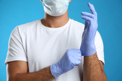 Photo of Man in protective face mask putting on medical gloves against blue background, closeup