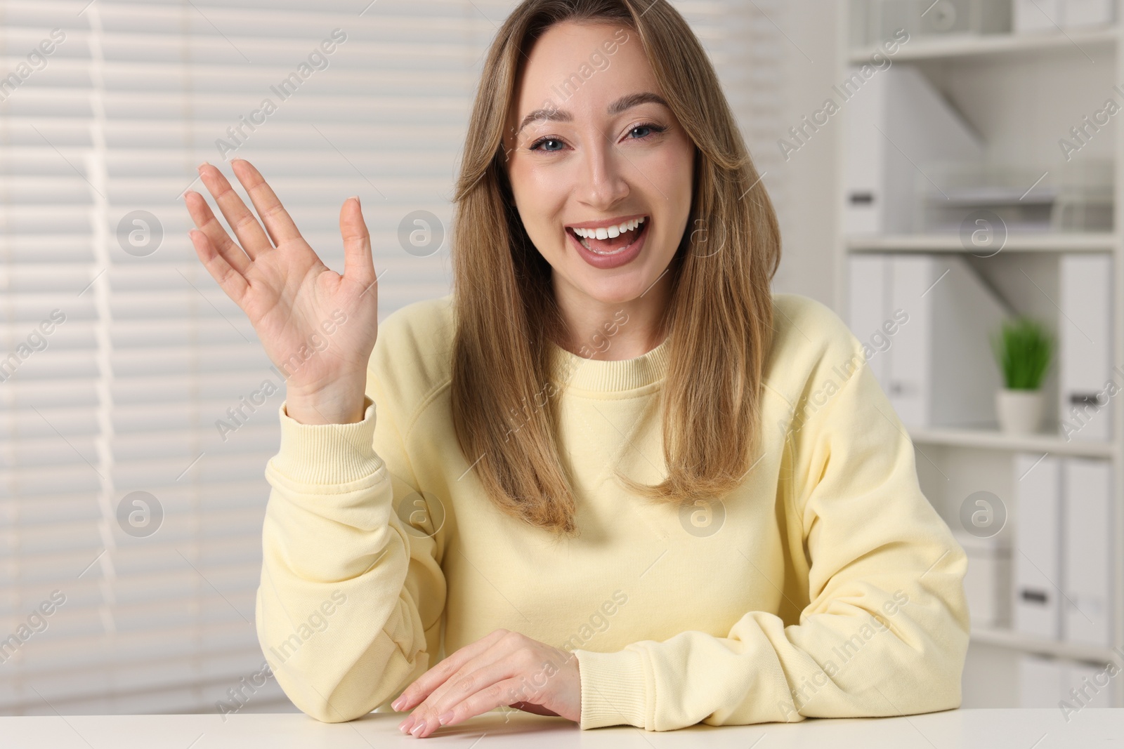 Photo of Young woman waving hello during video chat at white table indoors, view from webcam