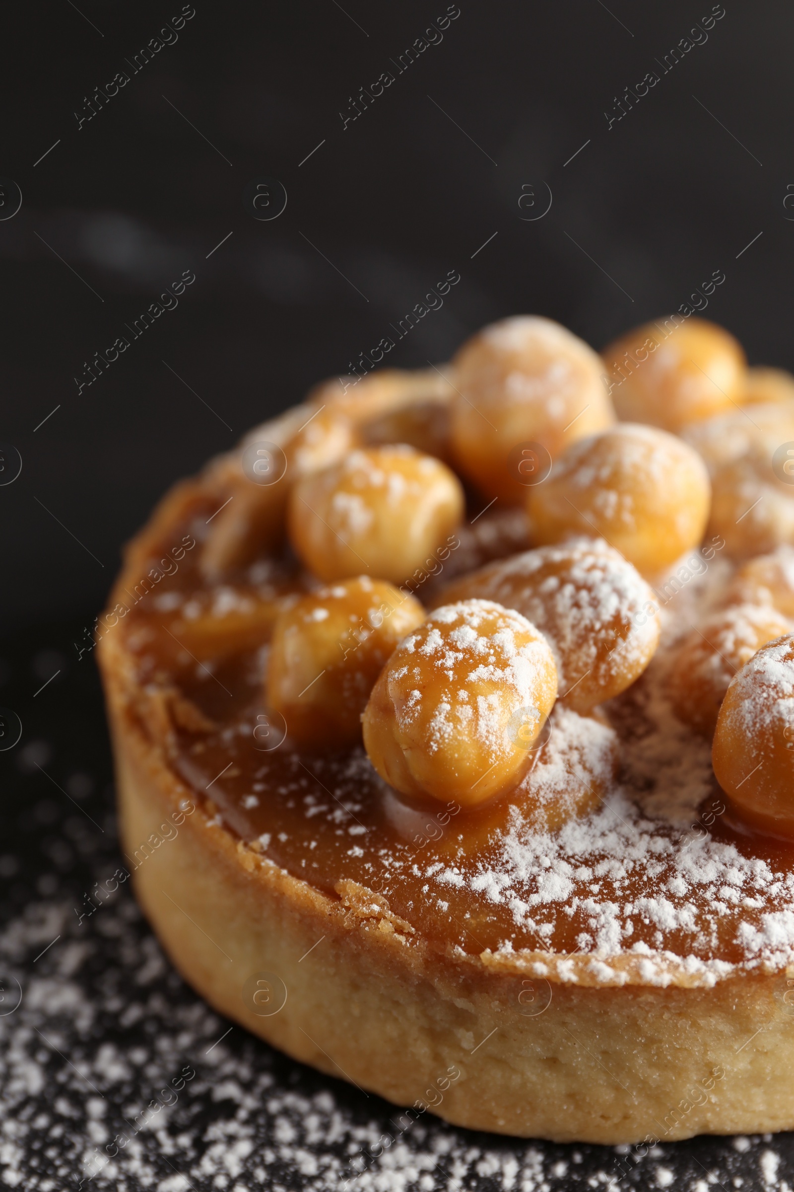 Photo of Delicious tart with hazelnuts, sweet caramel and powdered sugar on black table, closeup