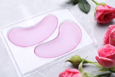 Photo of Package of under eye patches and rose flowers on light grey table. Cosmetic product